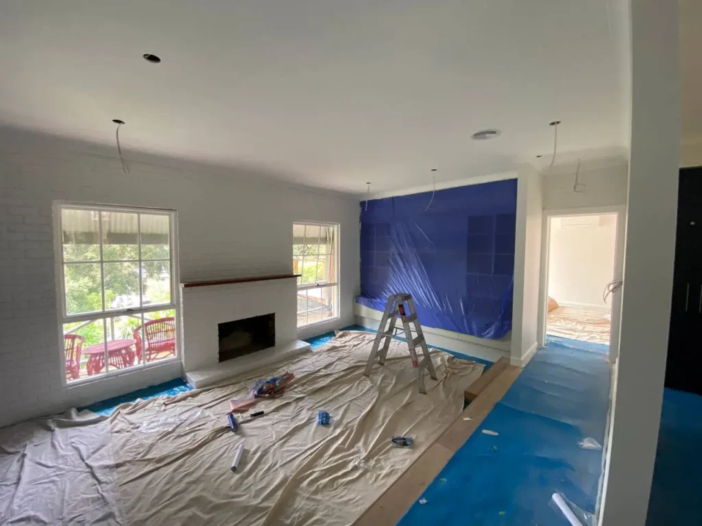 A living room interior under renovation with professional paint preparation, drop sheets, and protective blue film covering the floors.