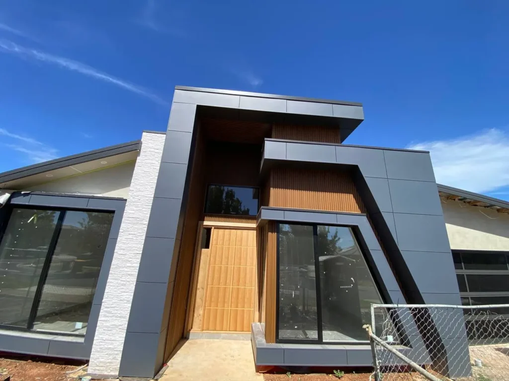 A newly finished modern house facade featuring charcoal grey architectural cladding panels, warm timber cladding accents, and painted render under a clear blue sky.